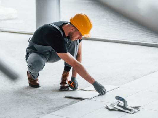 Handwerker mit gelbem Schutzhelm misst präzise Bodenfliesen aus, bevor sie verlegt werden.
