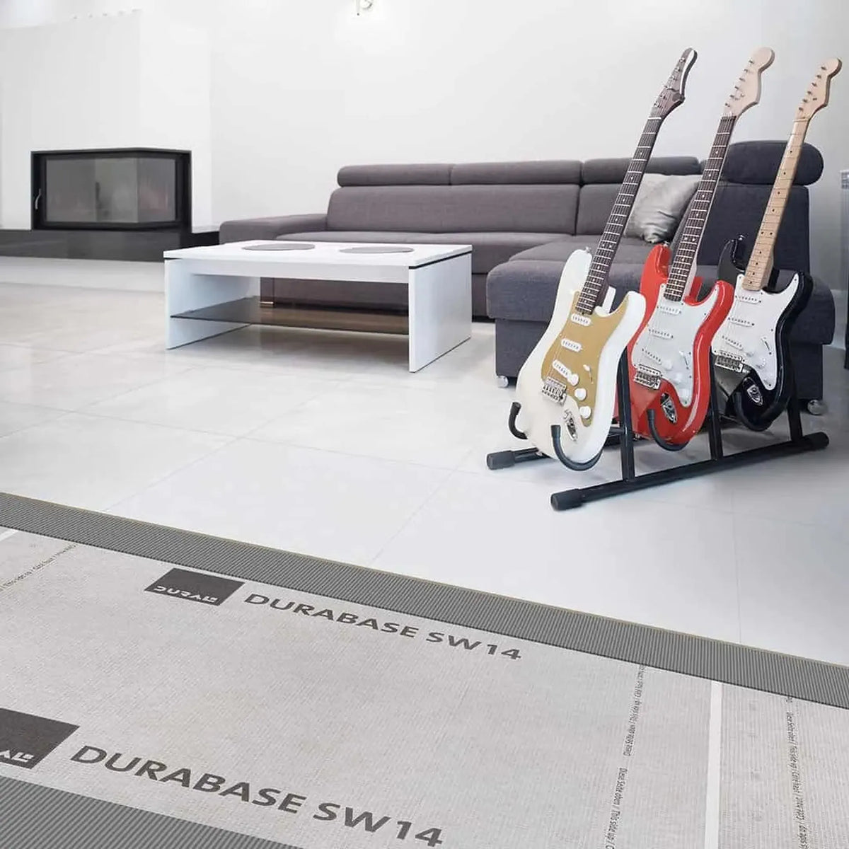 DURAL Trittschallmatte SW14 in einem Wohnzimmer unter weißen Fliesen verlegt. Zu sehen sind noch eine Couch, Tisch, Ofen und drei E-Gitarren