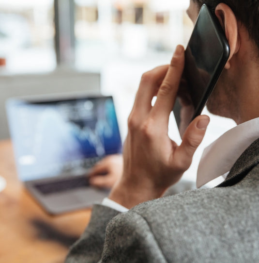 Geschäftsmann in grauem Anzug telefoniert mit dem Smartphone, während er auf einen Laptop mit Finanzdiagrammen blickt.