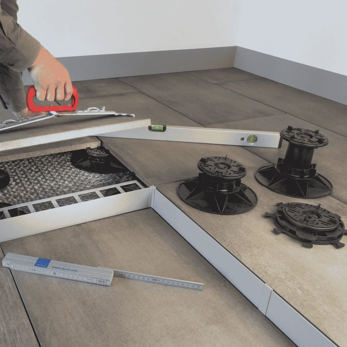 Ambientebild der 3 verschiedenen Stelzlagergrößen auf einer Terrasse. Der Handwerker benutzt eine Wasserwaage und einen Plattenheber zum Fliesenlegen.  #A0005386 #A0005387 #A0005388