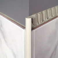 Profilé en quart de cercle en aluminium revêtu par poudrage couleur sable avec bord apparent arrondi, découpe triangulaire et aspect mat, posé à l'angle extérieur sous un carrelage clair.